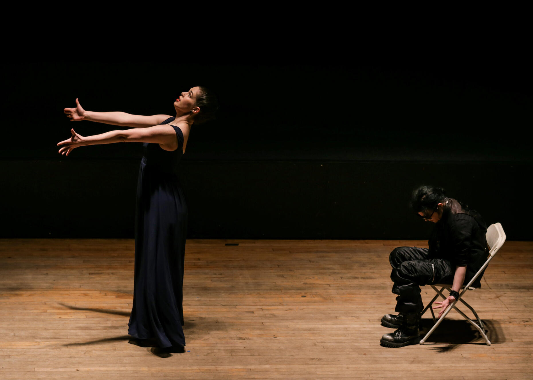A photograph of Miss Brents facing the left side of the stage, both arms outstretched and looking slightly upward. She wears a dark blue dress. Behind her, on the right side of the stage, her dance partner, wearing all black casual clothing, slumps in a fo